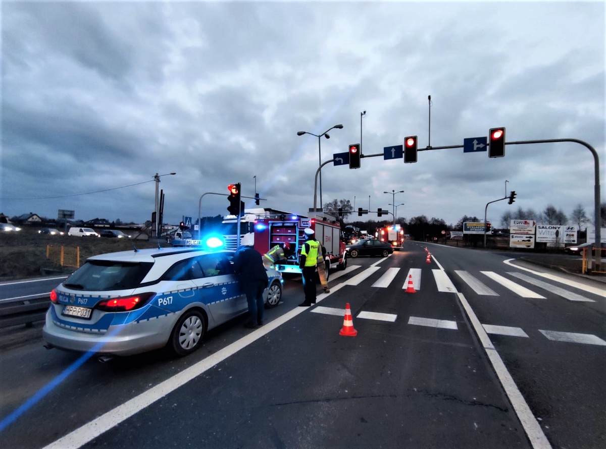 Zdjęcie kolorowe: skrzyżowanie świetlne, na pasie do lewoskrętu stoi oznakowany radiowóz policyjny, w tle widoczni policjanci i strażacy