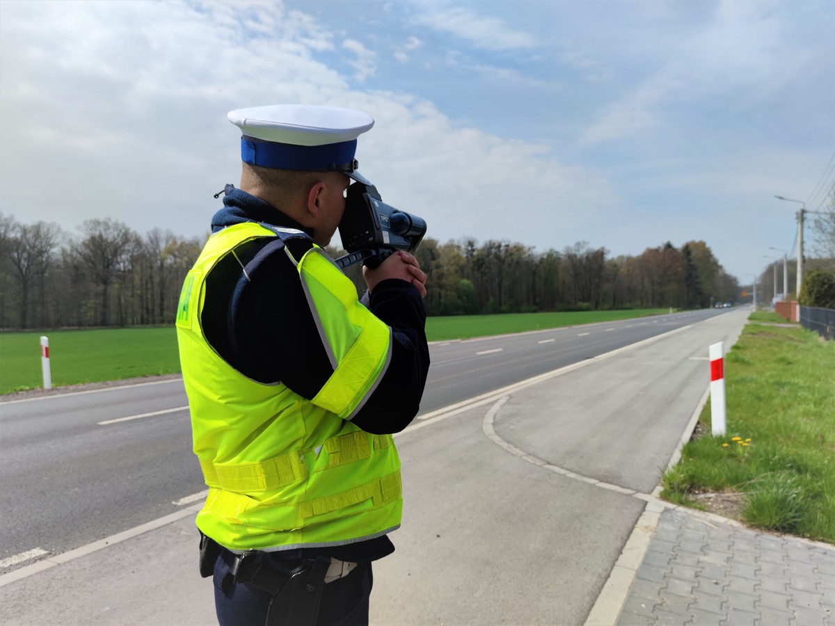 Zdjęcie kolorowe: umundurowany policjant ruchu drogowego podczas pomiaru prędkości pojazdów.