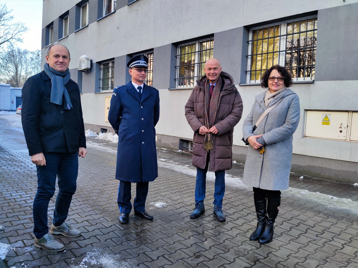 Zdjęcie przedstawia umundurowanego policjanta, kobietę i dwóch mężczyzn. Z tyłu widoczna komenda.