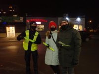 Widać dwie osoby , mężczyznę i kobietę, którzy trzymają w ręku odblaski. Obok nich stoi umundurowany policjant. Jest zmrok. W tle widać sklepy