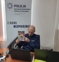 Umundurowany policjant w pomieszczeniu biurowym. Trzyma w ręku broszurę z napisem Narkotyki. Przed nim znajduje się laptop. W tle napis Komenda Powiatowa Policji w Pszczynie