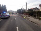 Zdjęcie przedstawia radiowóz policji ustawiony po lewej stronie drogi. W oddali znajduje się ambulans pogotowia. Na jezdni policjanci, strażacy i ratownicy medyczni. Po prawej stronie zniszczone kamienne ogrodzenie prywatnej posesji.