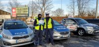 Dwóch umundurowanych policjantów na tle radiowozu znajdującego się na parkingu