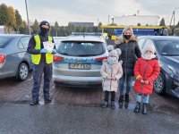 Pora dzienna. Kobieta z dwójką córek i umundurowany policjant na tle radiowozu. Wszyscy posiadaja elementy odblaskowe. Znajdują sie na parkingu