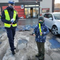 Pora dzienna. Umundurowany policjant z chłopcem który otrzymał opaskę odblaskową. Znajdują się na parkingu