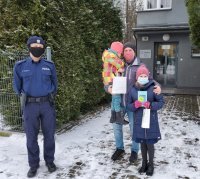 Umundurowany policjant w obecności dwóch dziewczynek i ich ojca stoją na tle komendy. Dziewczynka trzyma w ręku tablet. Pora dzienna, widoczne zaśnieżenia