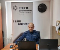 Umundurowany policjant siedzący przed biurkiem. Przed nim znajduje się komputer. Za policjantem znajduje się baner z napisem Komenda Powiatowa Policji w Pszczynie, Z Nami Bezpiecznie