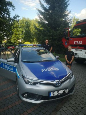 Zdjęcie kolorowe: na pierwszym planie radiowóz, obok stoi chłopiec, za nim policjant. W tle widoczny wóz straży pożarnej.