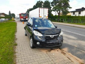 Zdjęcie kolorowe: na pierwszym planie uszkodzony czarny samochód osobowy, w tle przyjeżdżające pojazdy