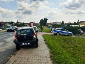 Zdjęcie kolorowe: na pierwszym planie uszkodzony czarny samochód osobowy, w tle przyjeżdżające pojazdy, wóz straży pożarnej