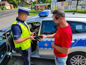 Zdjęcie kolorowe: policjant ruchu drogowego podczas kontroli drogowej obok mężczyzna w czerwonej koszulce opłaca mandat karny przy użyciu terminalu płatniczego