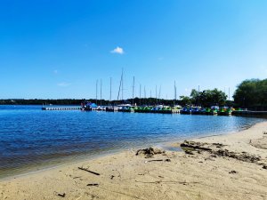 Zdjęcie kolorowe: na pierwszym planie plaża i woda, w tle żaglówki