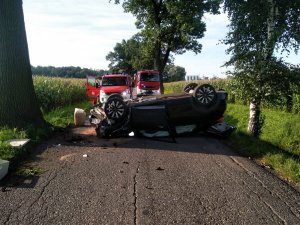 Zdjęcie kolorowe: na pierwszym planie widoczny samochód na dachu, w tle dwa wozy straży pożarnej