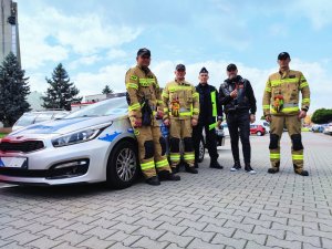 Zdjęcie kolorowe: trzech strażaków, policjant i mężczyzna w stroju motocyklowym z papryczką w dłoni na tle radiowozu.