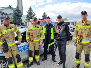 Zdjęcie kolorowe: trzech strażaków, policjant i mężczyzna w stroju motocyklowym z papryczką w dłoni na tle radiowozu.