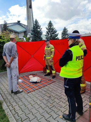 Zdjęcie kolorowe: strażak przy fantomie udziela instruktażu udzielania pierwszej pomocy, obok stoi wysoki mężczyzna oraz policjantka ruchu drogowego