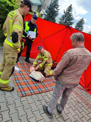 Zdjęcie kolorowe: strażak przy fantomie udziela instruktażu udzielania pierwszej pomocy, obok stoi mężczyzna oraz policjant