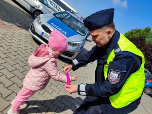 Zdjęcie kolorowe: policjant zakłada dziewczynce odblask, w tle radiowóz