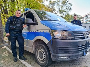 Zdjęcie kolorowe: dwóch umundurowanych policjantów stoi przy radiowozie