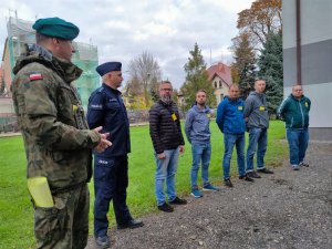 Zdjęcie kolorowe: widoczny umundurowany żołnierz, następnie policjant i kilku mężczyzn w szeregu. Wszyscy mają wzrok skierowany na mówiącego żołnierza.