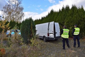 Zdjęcie kolorowe: na pierwszym planie widocznych dwóch umundurowanych policjantów stojących za białym samochodem typu bus. W tle widoczny radiowóz i umundurowany policjant.