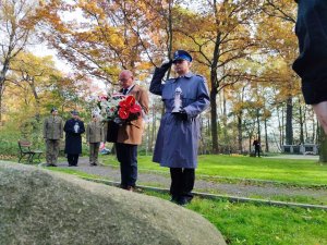 Zdjęcie kolorowe: umundurowany policjant oddaje honor, w rękach trzyma znicz. Obok stoi mężczyzna w eleganckim płaszczu i trzyma kwiaty.