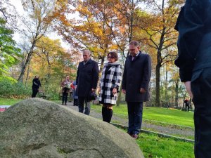 Zdjęcie kolorowe: trzy osoby widoczne na zdjęciu: mężczyzna, kobieta i mężczyzna, wszyscy mają spuszczone głowy oddając honor.