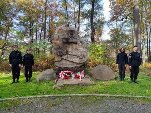 Zdjęcie kolorowe: na środku kamień pamiątkowy, z obu stron stoją po dwie młode osoby w mundurach