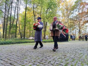 Zdjęcie kolorowe: umundurowany policjant idzie z kwiatami wraz z mężczyzną który niesie znicz