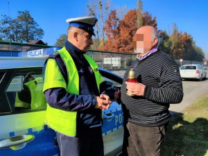 Zdjęcie kolorowe: policjant wręcza mężczyźnie znicz z kartką na której widnieje napis Zapal tym, co nie dojechali... W tle widoczny radiowóz.