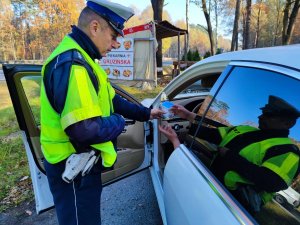 Zdjęcie kolorowe: policjant stoi obok samochodu osobowego, wewnątrz kierowca. Mundurowy wręcza znicz z kartką na której widnieje napis Zapal tym, co nie dojechali...