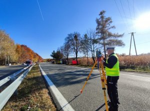 Zdjęcie kolorowe: policjant ruchu drogowego podczas czynności na miejscu wypadku drogowego. W tle widoczny wywrócony pojazd.