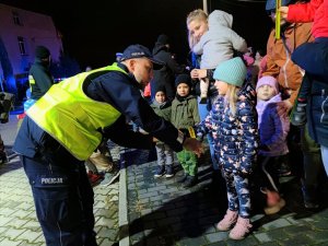 Zdjęcie kolorowe: umundurowany policjant zakłada opaskę odblaskową dziewczynce na rękę. W tle widoczne inne zebrane osoby.