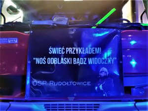 Zdjęcie kolorowe: na zdjęciu widoczny plakat, na którym jest strażak i widnieje napis o treści Świeć przykładem! Noś odblaski, bądź widoczny. OSP Rudołtowice. Całość zawieszona na wozie straży pożarnej.