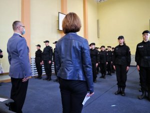 Zdjęcie kolorowe: na pierwszym planie kobieta i umundurowany policjant którzy stoją tyłem. Przed nimi grupa młodzieży w czarnych mundurach.