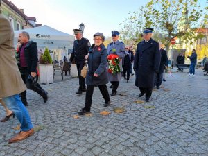 Zdjęcie kolorowe: widoczny pochód. Na pierwszym planie starsza pani uśmiecha się do obiektywu, w tle umundurowany policjant, strażak miejski i straży pożarnej oraz inni uczestnicy