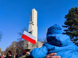 Zdjęcie kolorowe: małe dziecko siedzi na barkach mężczyzny z flagą biało-czerwoną w ręce, w tle widoczny pomnik