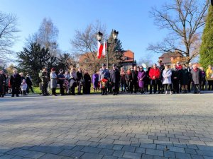 Zdjęcie kolorowe: umundurowany policjant trzyma w ręku kwiaty, stoi obok przedstawiciela straży pożarnej