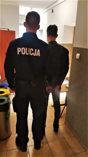 Zdjęcie kolorowe: umundurowany policjant stoi w korytarzu z zatrzymanym mężczyzną