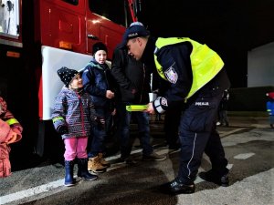 Zdjęcie kolorowe: umundurowany policjant wręcza uśmiechniętej dziewczynce odblaski, w tle widoczni inni uczestnicy