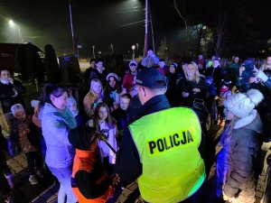 Zdjęcie kolorowe: na pierwszym planie widoczny umundurowany policjant, który stoi naprzeciw grupy ludzi