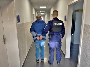 Zdjęcie kolorowe: umundurowany policjant stoi w korytarzu trzymając zatrzymanego, który założone kajdanki z tyłu