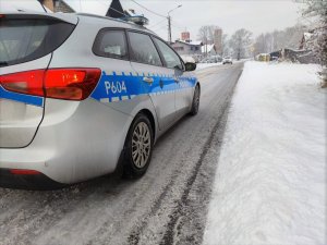 Zdjęcie kolorowe: oznakowany policyjny radiowóz w zimowej aurze. W tle widoczny inny pojazd