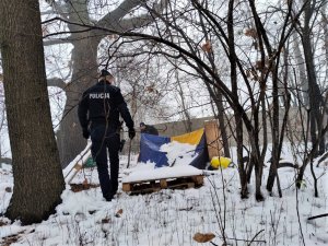 Zdjęcie kolorowe: na pierwszym planie umundurowany policjant, w tle widoczny namiot. Na ziemi widoczny śnieg