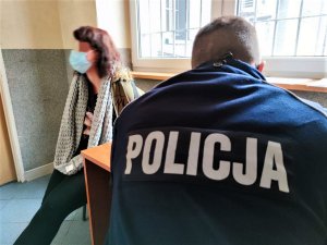 Zdjęcie kolorowe: na pierwszym planie umundurowany policjant, który siedzi tyłem. W tle kobieta z maską na twarzy, siedzi.