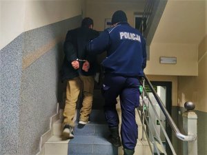 Zdjęcie kolorowe: umundurowany policjant prowadzi po schodach zatrzymanego mężczyznę