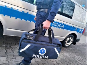 Zdjęcie kolorowe: umundurowany policjant z torbą pierwszej pomocy na tle oznakowanego radiowozu