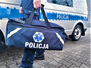 Zdjęcie kolorowe: umundurowany policjant z torbą pierwszej pomocy na tle oznakowanego radiowozu