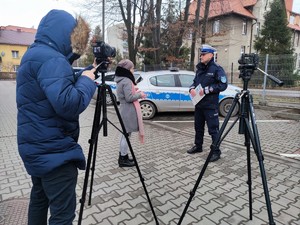 Zdjęcie kolorowe: widoczne dwa aparaty na statywach, za jednym stoi mężczyzna z kapturem na głowie. Przed obiektywem widoczny umundurowany policjant ruchu drogowego i kobieta. W tle policyjne radiowozy.