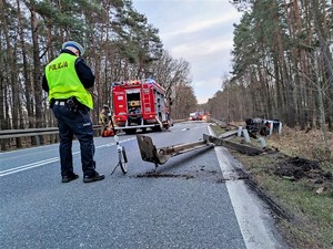 Zdjęcie kolorowe: umundurowany policjant ruchu drogowego podczas czynności na miejscu zdarzenia drogowego. Obok niego stoi wózek do pomiaru. W tle widoczny wóz straży pożarnej.